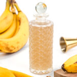 A textured glass bottle with a round stopper is surrounded by bananas, a wooden board, a yellow cloth, and a jigger, set against a white background.