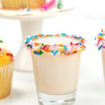 A shot glass filled with a creamy birthday cake shot, rimmed with colorful sprinkles. Cupcakes sit in the background, while candles flicker nearby.