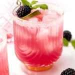 A pink cocktail in a ridged glass, garnished with lime, mint, and a blackberry. The rim is sugared, with blackberries and mint scattered on a marble surface.