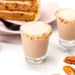 Two cream-colored shots with nut-crusted rims on a marble surface, accompanied by scattered pecans, a slice of layered cake on a pink plate, a black fork, and a golden jigger.