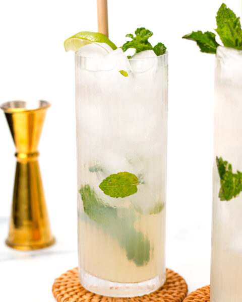 A tall glass of iced mojito with mint leaves and a lime wedge, placed on a wicker coaster. A gold jigger is in the background.