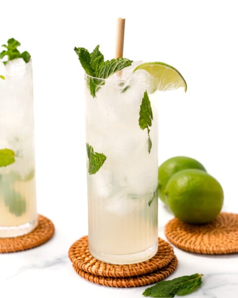 Tall glass of iced mojito with mint leaves, lime wedge, and a straw, placed on a woven coaster. Two limes in the background on a white surface.