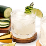 A glass of iced cocktail garnished with a cucumber ribbon and lime slice, placed on a wooden coaster. Sliced cucumber, lime wedge, and a metal jigger are nearby.