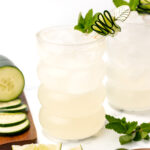 Two glasses of a refreshing cocktail with ice, each garnished with a cucumber spiral and mint. Sliced cucumber and lime wedges are arranged on a wooden board nearby.