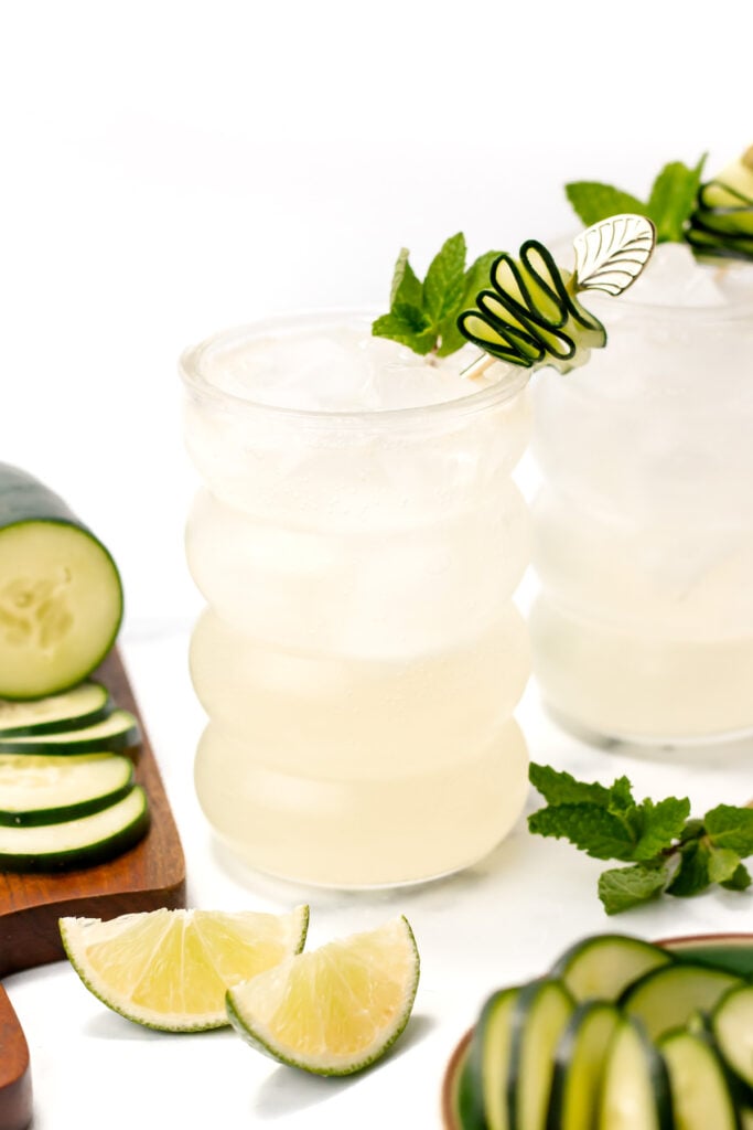 Two glasses of a refreshing cocktail with ice, each garnished with a cucumber spiral and mint. Sliced cucumber and lime wedges are arranged on a wooden board nearby.