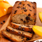 Sliced chocolate chip banana bread on a wooden board, surrounded by bananas and a small bowl of chocolate chips.