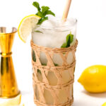 A tall glass with ice, mint, and a lemon slice on the rim, wrapped in wicker. A gold jigger and whole lemons are nearby on a white surface.