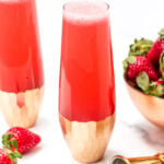 Two glasses filled with a red sparkling drink, partially obstructed by a gold ombre base, next to fresh strawberries and a metal jigger.