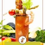 A Bloody Mary cocktail in a tall glass, garnished with shrimp, bacon, celery, olives, asparagus, pickles, a cherry tomato, and lime, with vegetables and herbs in the background.