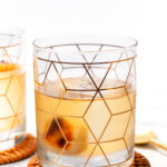 A glass with geometric patterns contains an amber-colored drink and an ice sphere, placed on a woven coaster.