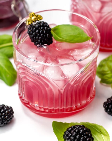 A glass of pink blackberry cocktail with ice, garnished with a blackberry and basil leaves, surrounded by fresh blackberries and basil.