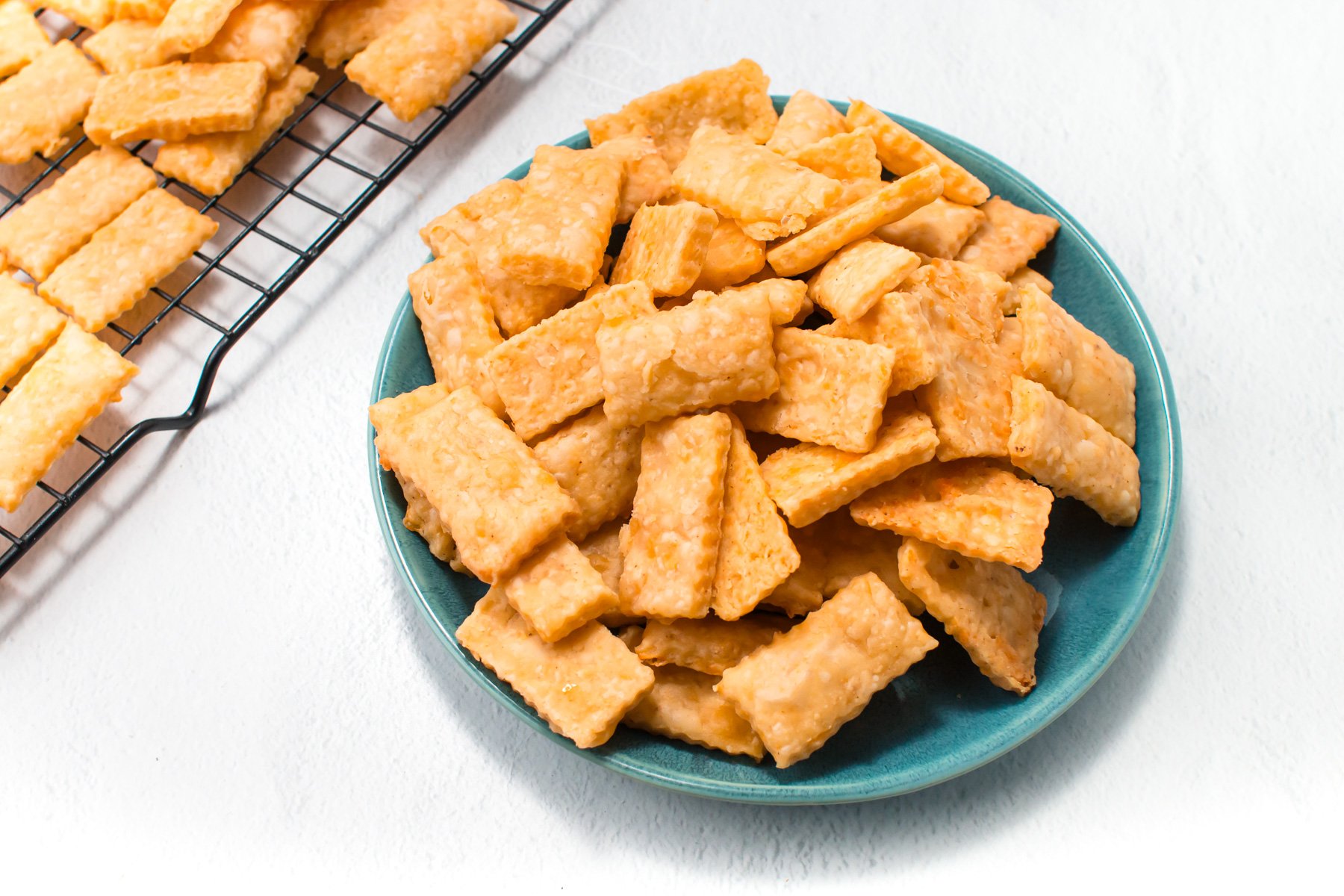A teal plate filled with square and rectangular cheese crackers on a white surface, next to a cooling rack with more crackers.
