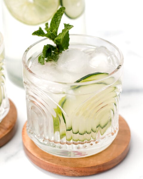 A glass of iced beverage garnished with mint and cucumber slices, placed on a wooden coaster.