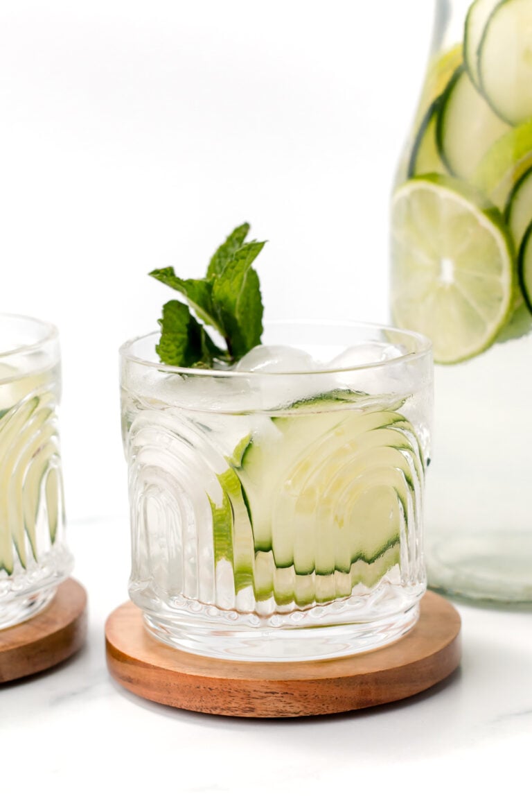 A glass of ice water with cucumber slices and mint on a wooden coaster, with a pitcher of water containing cucumber and lime slices in the background.