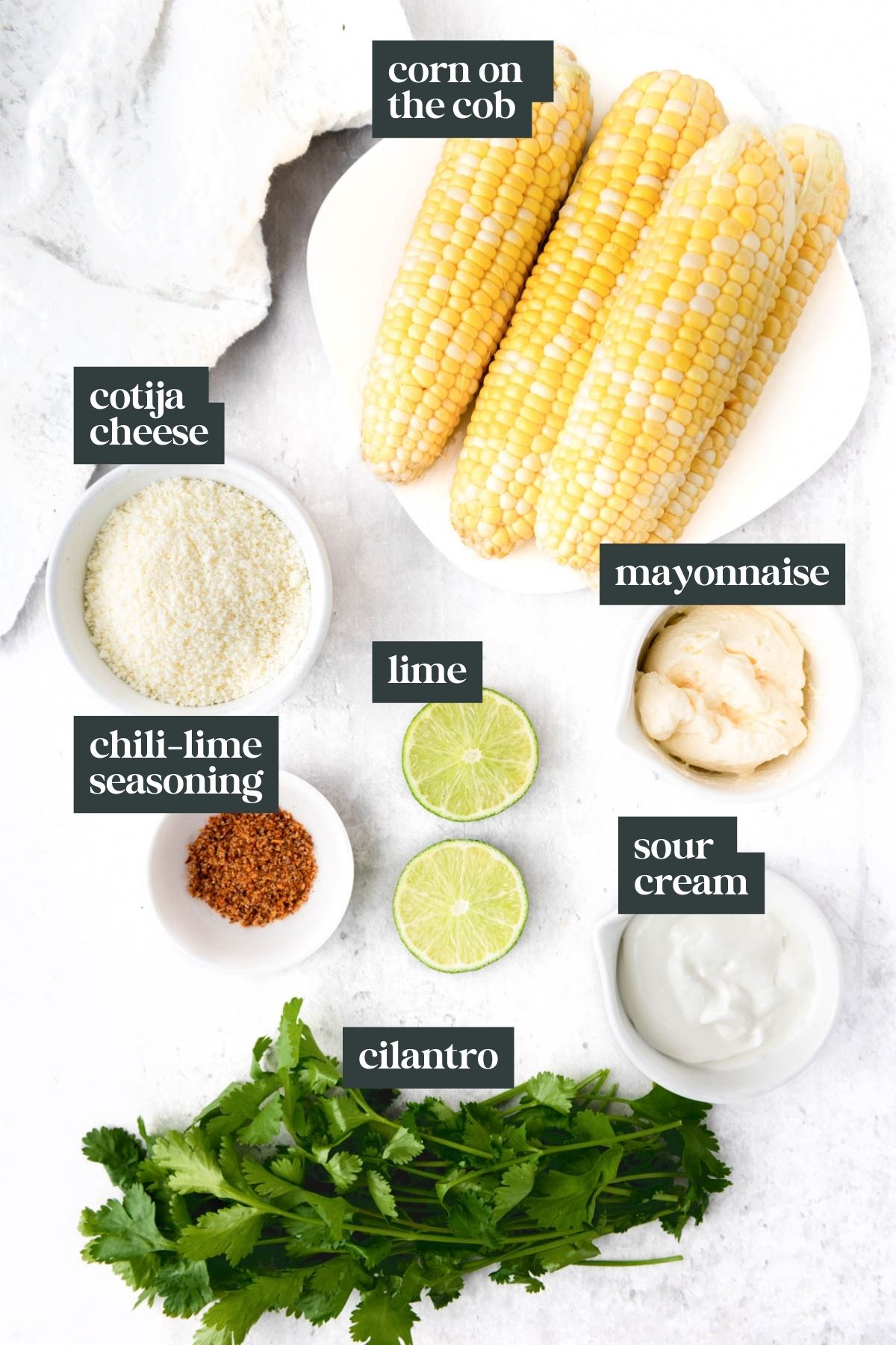 Corn on the cob with cotija cheese, mayonnaise, sour cream, chili-lime seasoning, lime halves, and cilantro arranged on a white surface.