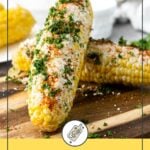 Two cobs of Mexican street corn topped with cheese and herbs on a wooden board, with seasoning scattered around.