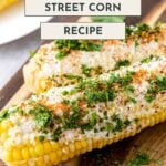 Two ears of Mexican street corn topped with cheese, chili powder, and cilantro on a wooden board.