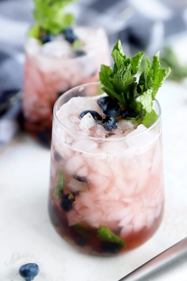 A glass of crushed ice cocktail garnished with fresh mint leaves and blueberries, with another similar drink in the background.