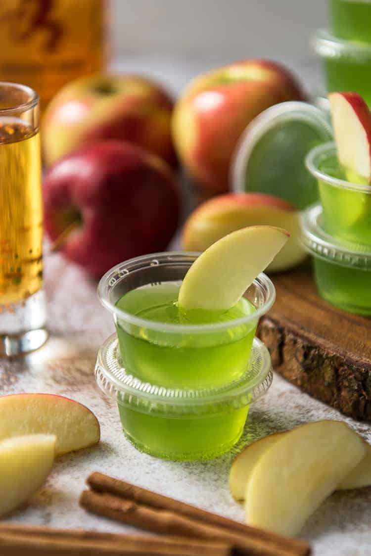 Small plastic cups filled with green gelatin, each topped with an apple slice, surrounded by apples, apple slices, and cinnamon sticks.