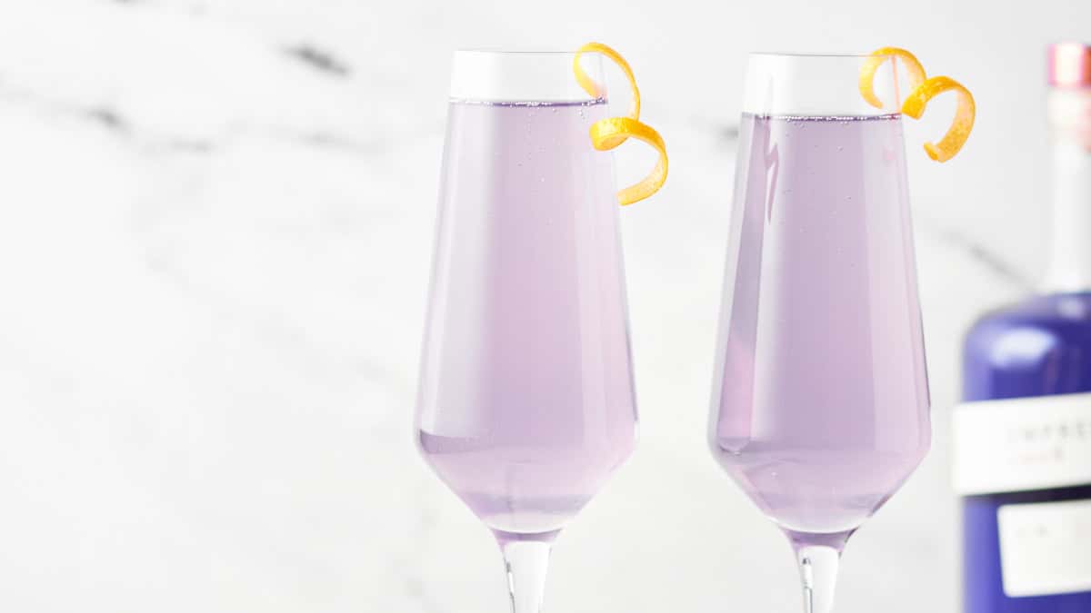 Two champagne flutes filled with a light purple cocktail, each garnished with a curled orange peel; a bottle is partially visible in the background.