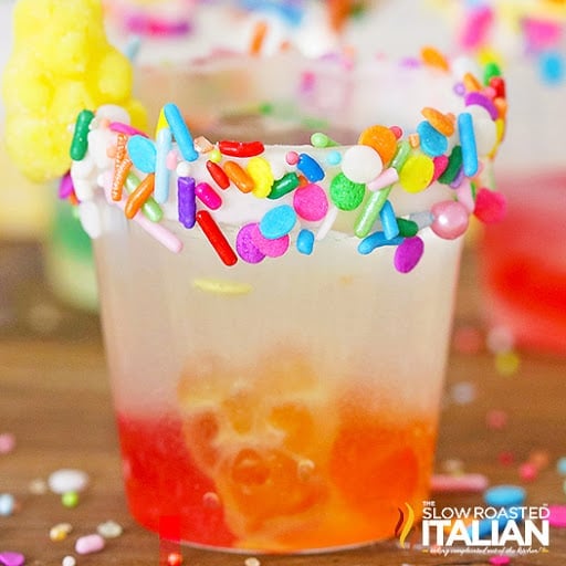 A clear glass with a colorful sprinkle-coated rim contains a layered red and yellow drink, garnished with a yellow candy.
