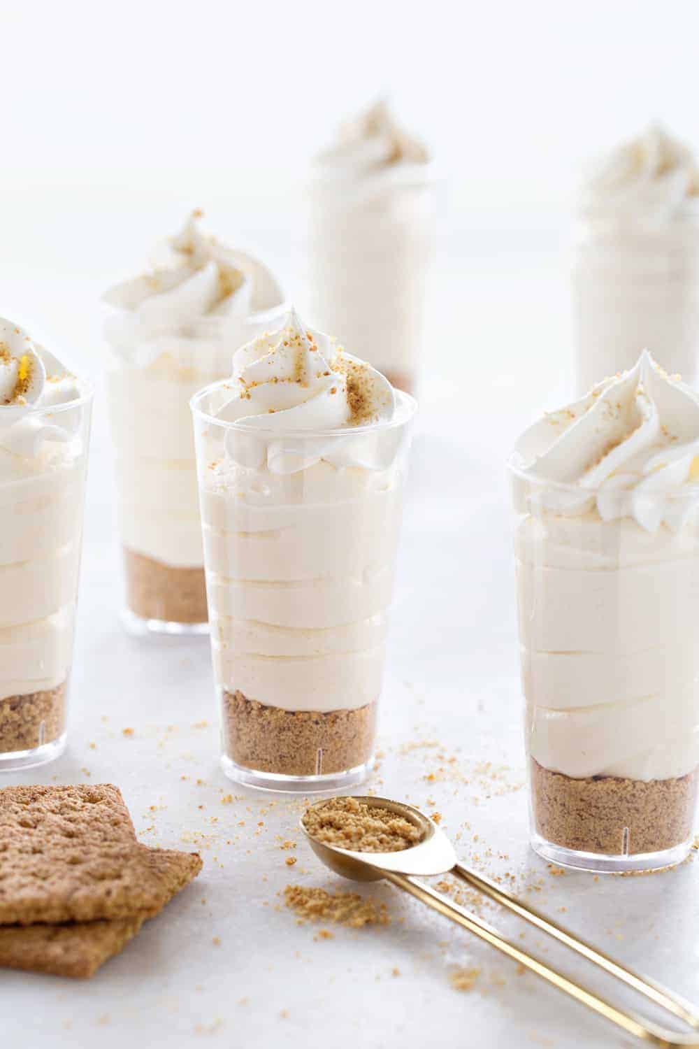 Several small clear cups filled with layered graham cracker crumbs and creamy cheesecake filling, topped with whipped cream, are arranged on a white surface with graham crackers nearby.