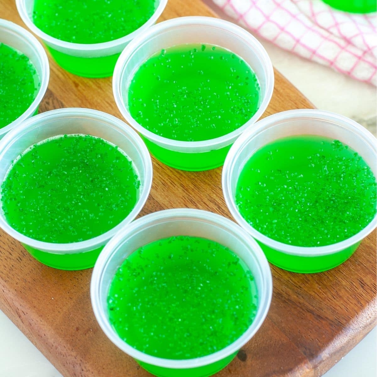 Six small clear plastic cups filled with bright green gelatin are arranged on a wooden surface. Some cups have a sprinkling of green sugar on top.