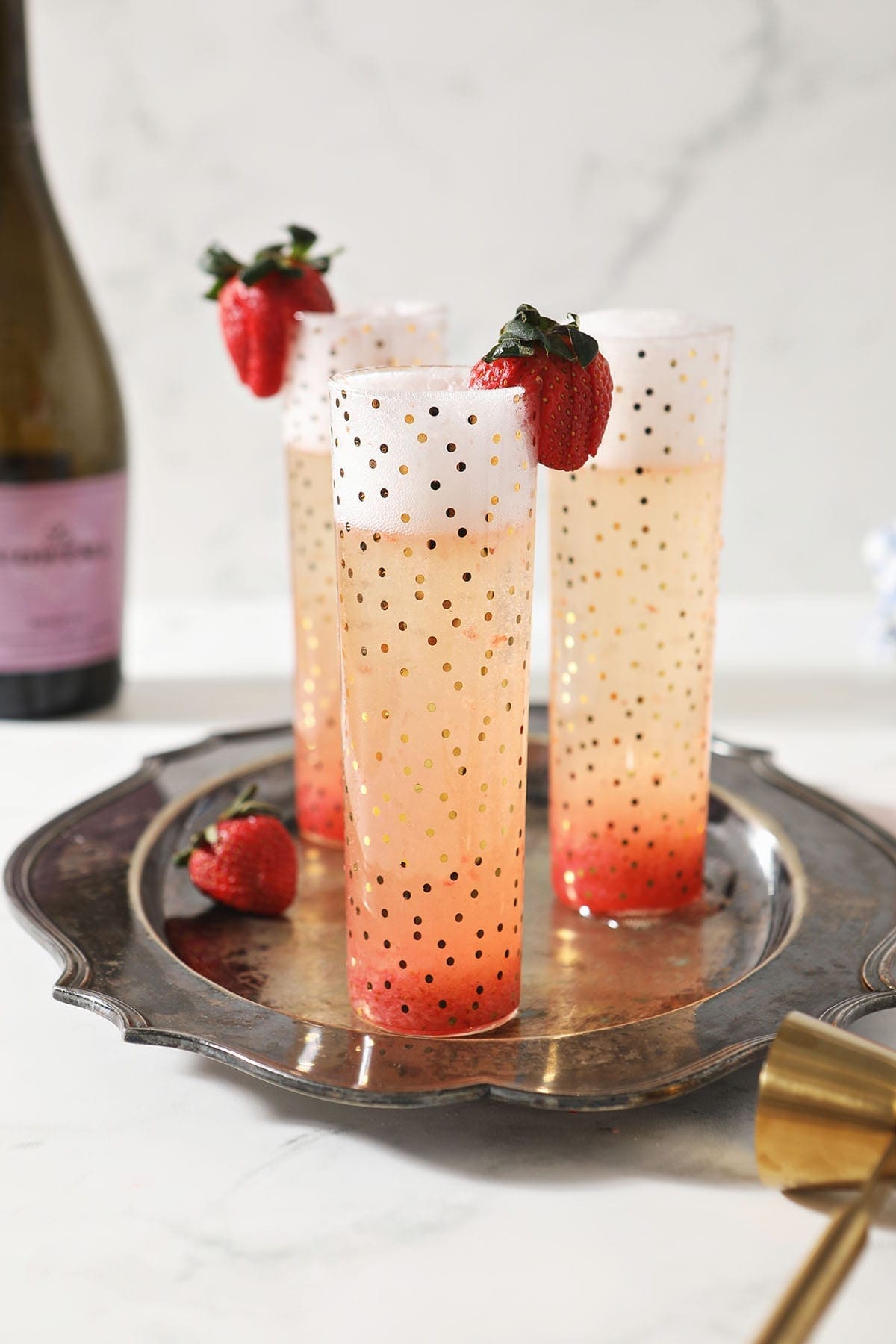 Three tall glasses with a pink sparkling drink, garnished with strawberries, sit on a silver tray; a wine bottle and gold jigger are in the background.