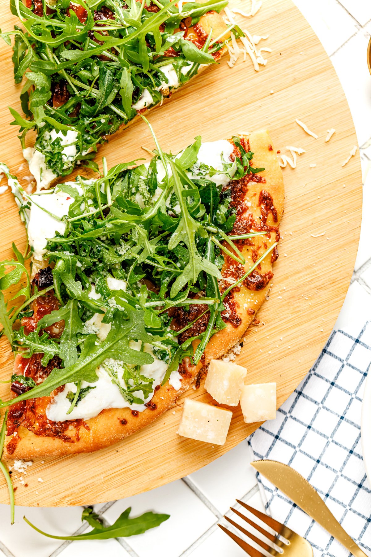 A sliced pizza topped with arugula and cheese sits on a wooden board, with Parmesan cubes and utensils nearby.