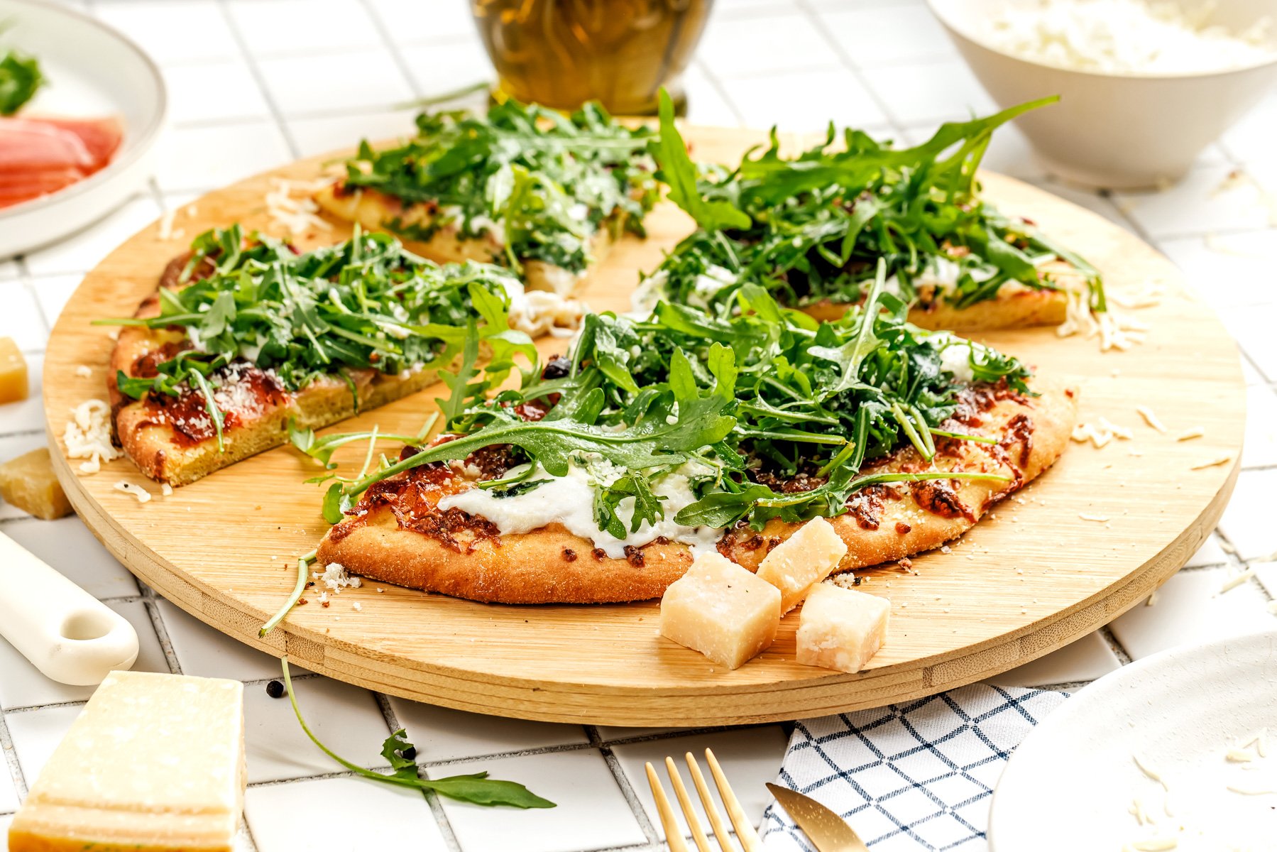 A sliced pizza topped with arugula, cheese, and tomato sauce sits on a wooden board, surrounded by pieces of cheese and a few kitchen items.