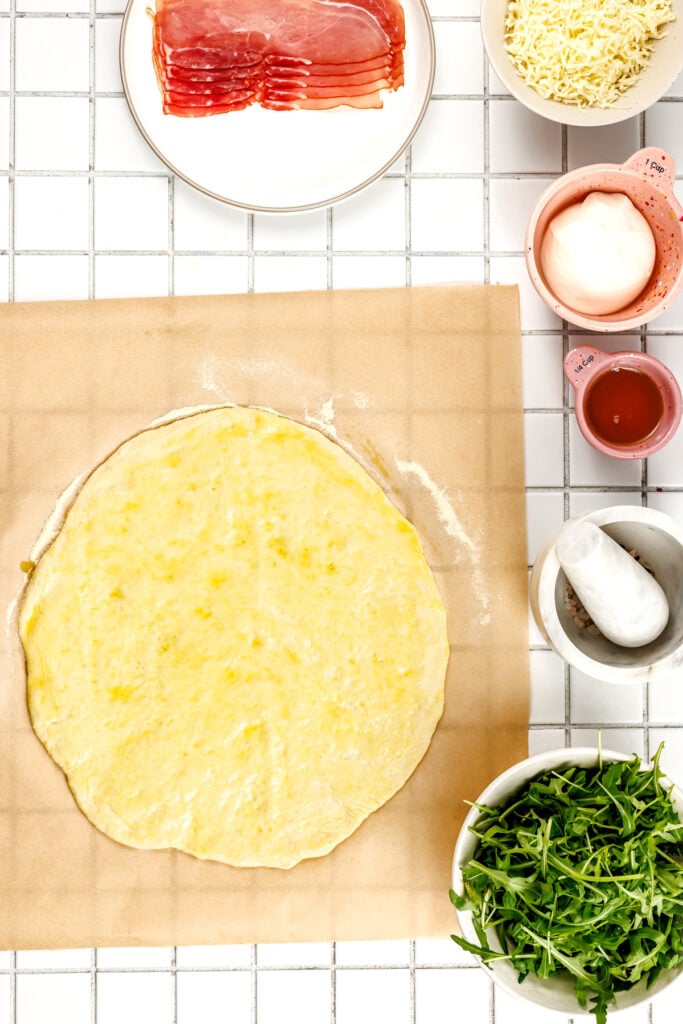 A rolled-out pizza dough on parchment paper is surrounded by bowls of arugula, mozzarella, sliced prosciutto, flour, tomato sauce, and a mortar and pestle.