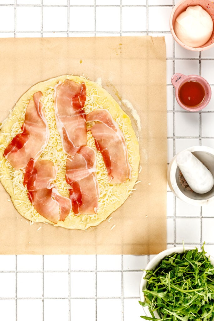 Uncooked pizza topped with cheese and prosciutto on parchment paper, surrounded by bowls of arugula, sauce, seasoning, and mayonnaise on a tiled surface.