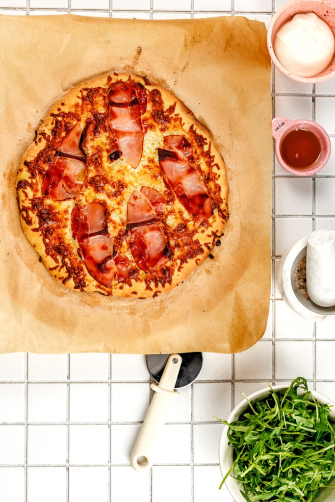 A baked pizza with ham on parchment paper next to a pizza cutter, a bowl of arugula, and small dishes of sauce and cream on a white tiled surface.