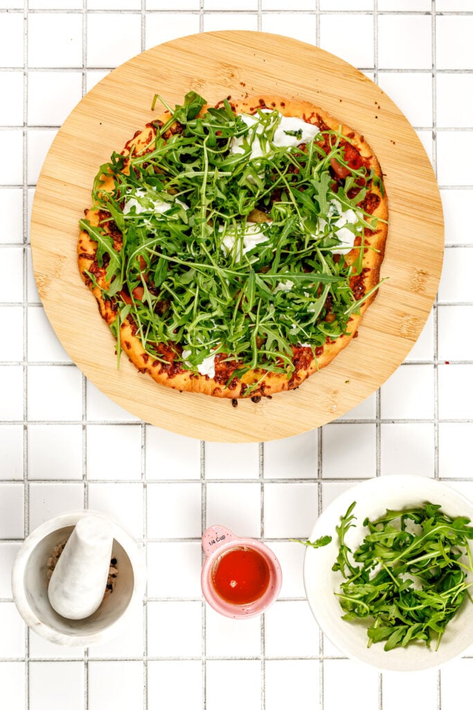 A pizza topped with arugula on a wooden board is placed on a white tiled surface, surrounded by a mortar and pestle, a small pitcher, and a bowl of arugula.