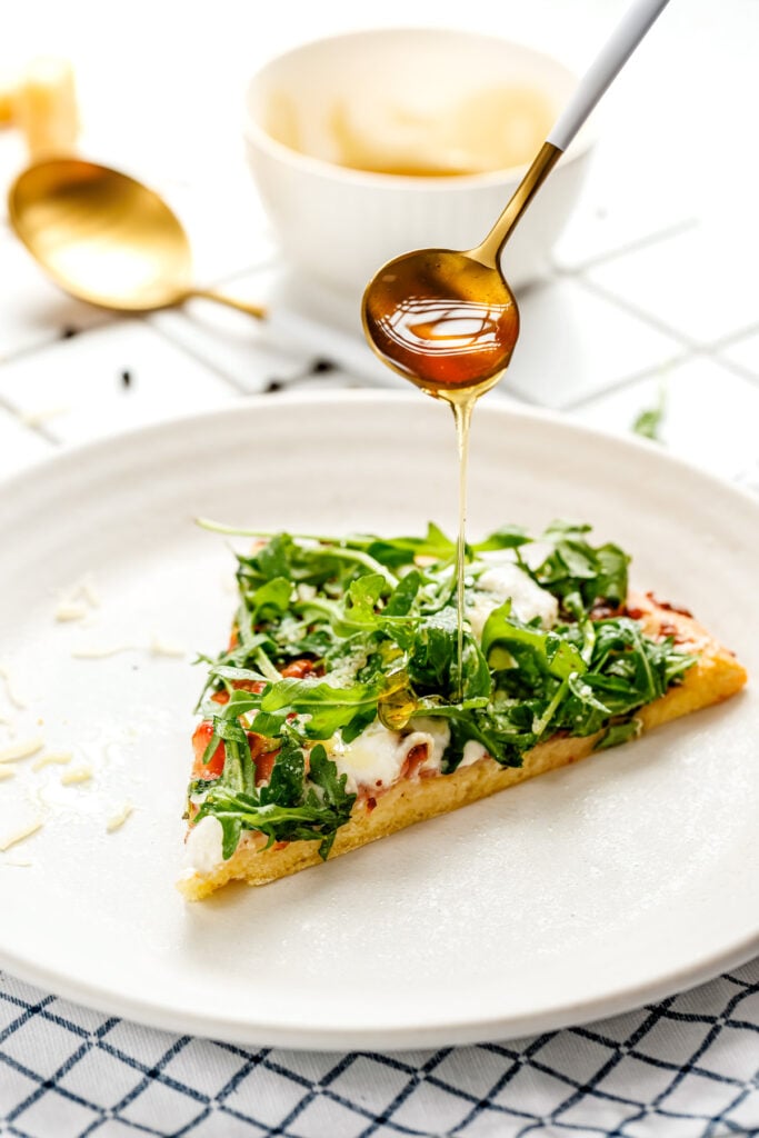 A spoon drizzles honey over a slice of pizza topped with arugula and cheese, placed on a white plate with a gold spoon and bowl in the background.