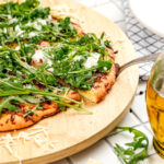 A pizza topped with arugula and cheese sits on a wooden board, with one slice being lifted by a metal spatula.