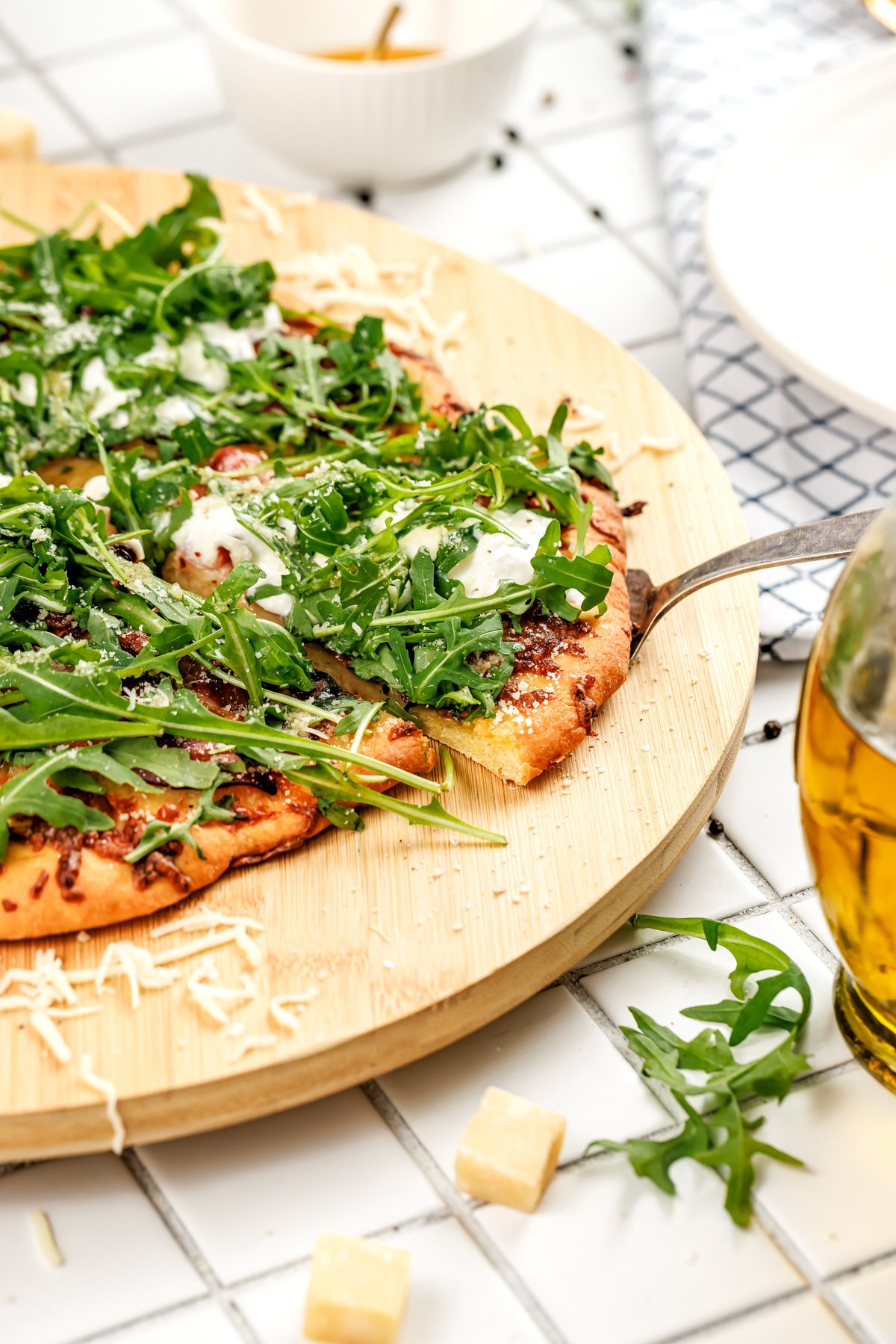 A pizza topped with arugula and cheese sits on a wooden board, with one slice being lifted by a metal spatula.