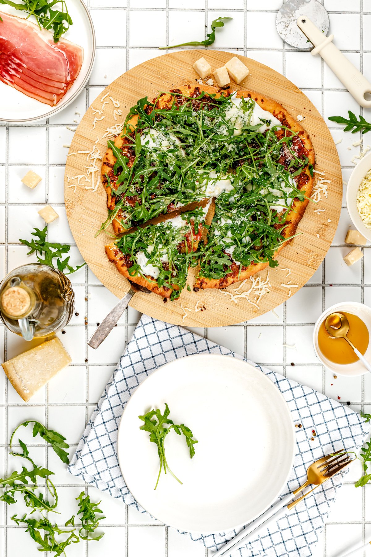 A pizza topped with arugula and cheese sits on a wooden board, surrounded by plates, utensils, parmesan cheese, olive oil, and prosciutto on a tiled surface.