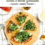A wooden board with sliced prosciutto and burrata pizza topped with arugula, surrounded by cheese, plates, and utensils on a white surface.