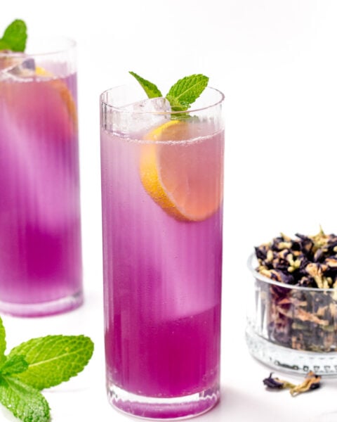 A tall glass of purple iced drink garnished with a lemon slice and mint, with a second glass, fresh mint, and a small bowl of dried flowers nearby.