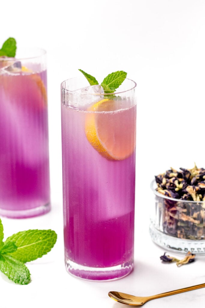 A tall glass of purple iced drink garnished with a lemon slice and mint, with a second glass, fresh mint, and a small bowl of dried flowers nearby.