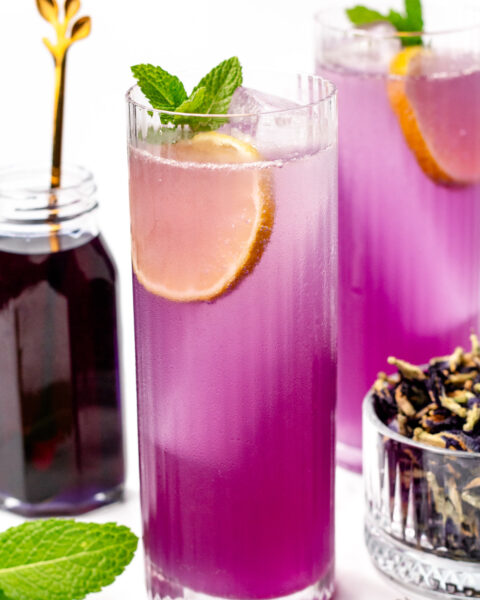 A tall glass of purple iced drink garnished with a lemon slice and mint, next to a jar of dark liquid, a bowl of dried flowers, and fresh mint leaves.