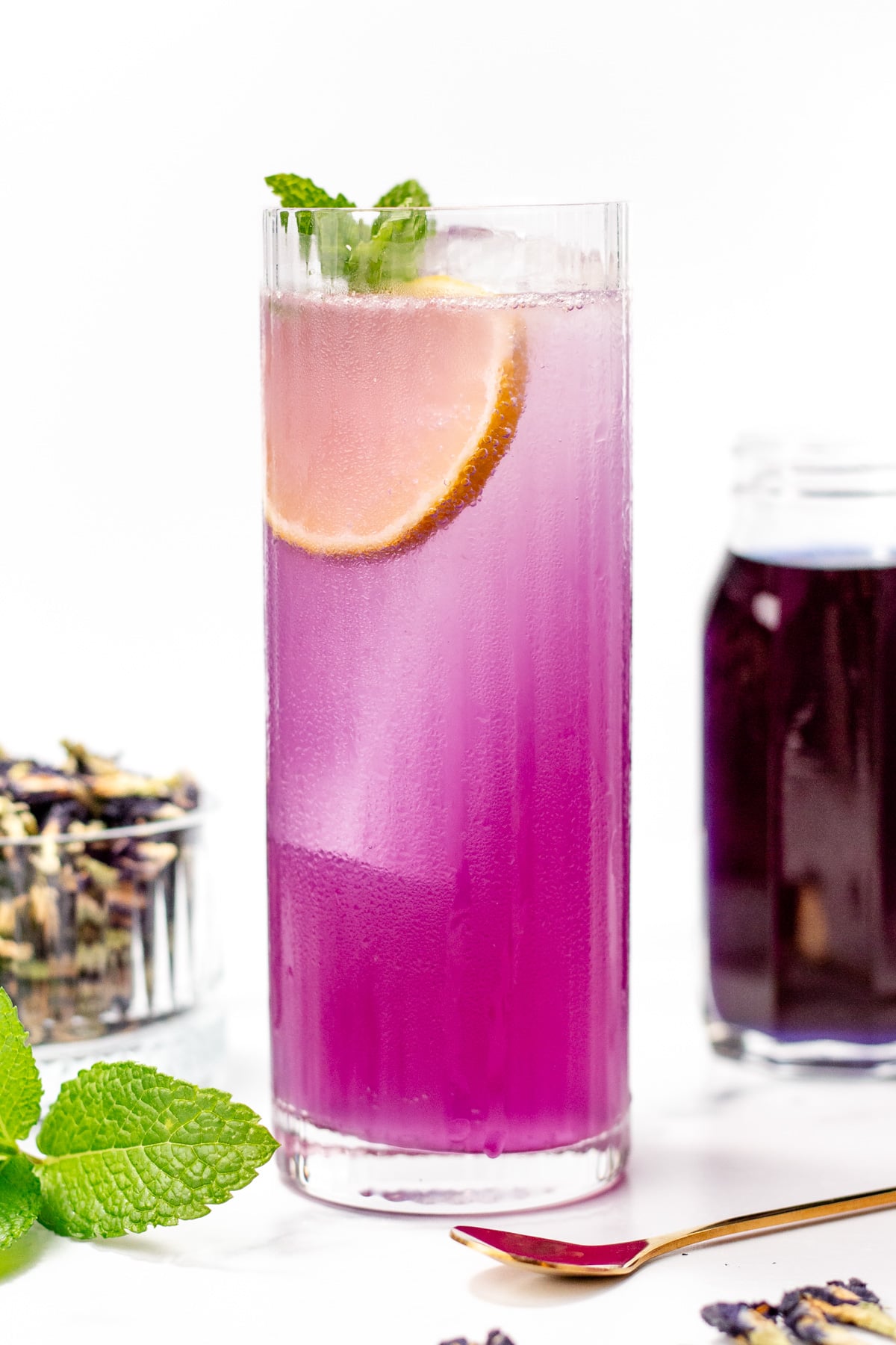 A tall glass of purple iced beverage garnished with a lemon slice and mint, with a jar of dark liquid and a bowl of dried flowers nearby.