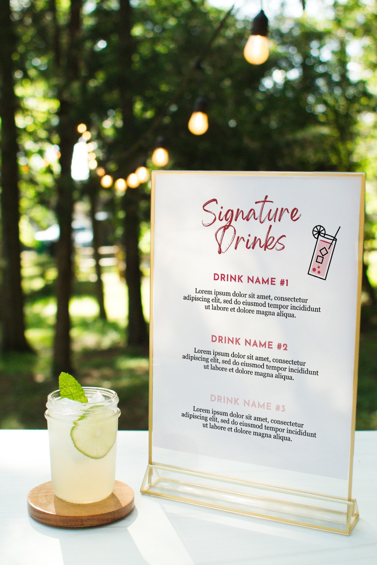 A glass of lemonade on a coaster sits next to a printed menu titled "Signature Drinks" on an outdoor table with string lights and trees in the background.