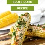 Two ears of Mexican elote corn topped with cheese, herbs, and spices are placed on a wooden board, with more corn and a white bowl in the background. Text reads “Mexican Elote Corn Recipe.”.