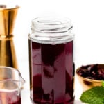 A glass jar filled with dark red hibiscus syrup sits on a white surface, surrounded by dried hibiscus petals, a metal jigger, and a bowl with more petals.