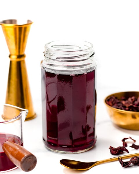 A jar filled with dark red liquid sits on a white surface, surrounded by a measuring jigger, a gold spoon, and a small bowl of dried hibiscus petals.