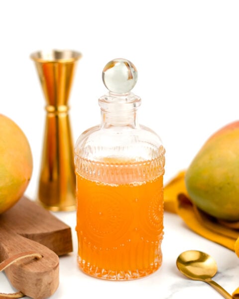 A glass decanter filled with orange liquid is surrounded by a jigger, a spoon, two mangoes, and a wooden cutting board on a white surface.
