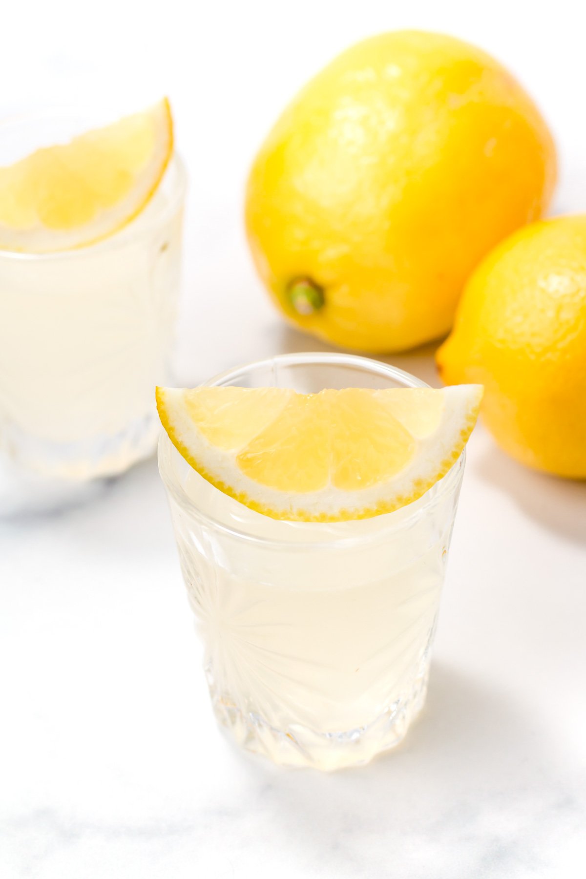 A glass of lemon juice garnished with a lemon wedge is in the foreground, with two whole lemons and another glass in the background on a white surface.