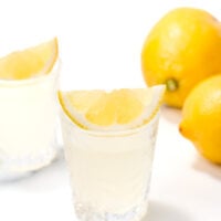 A glass of lemon juice with a lemon wedge on the rim, another glass, and whole lemons in the background on a white surface.